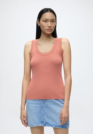Femme aux longs cheveux foncés portant un haut corail sans manches et une jupe en jean bleu clair, debout devant un fond blanc uni.