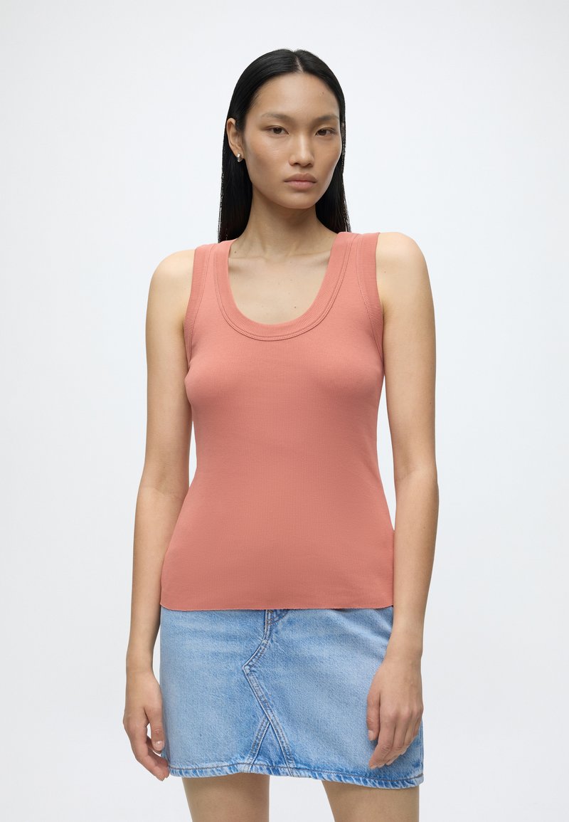 Femme aux longs cheveux foncés portant un haut corail sans manches et une jupe en jean bleu clair, debout devant un fond blanc uni.