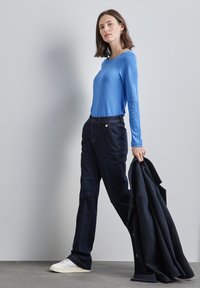 Ljusblå långärmad tröja, mörka denimbyxor med knapppdetaljer, håller en svart jacka, vita sneakers, står mot en grå bakgrund.