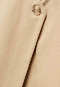 Tessuto beige con una trama strutturata, caratterizzato da un grande bottone rotondo sulla cucitura. Il tessuto è liscio con cuciture ben visibili.