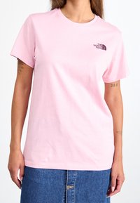 T-shirt in cotone rosa chiaro con maniche corte, scollatura rotonda e un piccolo logo nero sul lato sinistro del petto. Texture liscia, design casual.