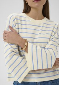 Pull à rayures jaune pâle avec des lignes bleues, doté de manches cloche et d'une coupe décontractée. Accessoirisé avec un bracelet doré.