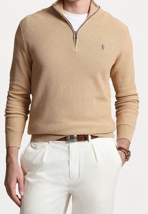 Mann trägt beigen Pullover mit Viertelreißverschluss über weißem Hemd, weiße plissierte Hose, braunen Gürtel und Armbanduhr am linken Handgelenk. Hände in den Taschen.