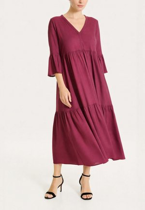 Donna che indossa un vestito midi fluido color bordeaux profondo con maniche a campana 3/4 e sandali neri con cinturino alla caviglia e tacco alto su uno sfondo chiaro.