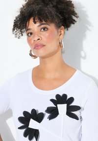 T-shirt bianco con due applicazioni di fiori neri con dettagli bianchi. La maglietta ha un collo rotondo ed è realizzata in tessuto morbido.