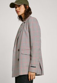 Blazer a quadretti nei toni beige, blu e rosso. Presenta un design doppiopetto, rever a punta, tasche frontali e spacchi laterali.