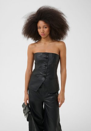 Femme aux grands cheveux bouclés portant un top noir en cuir sans bretelles avec des boutons et un pantalon en cuir noir, tenant une pochette noire.