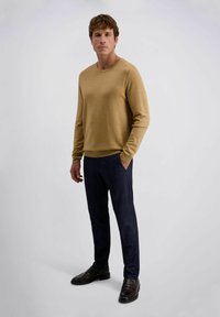 Mann steht mit entspannten Händen, trägt einen kamelfarbenen Pullover, dunkelmarineblaue Hosen und dunkelbraune Slipper vor einem einfarbigen hellen Hintergrund.