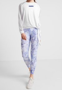 Donna che indossa una maglietta bianca a maniche lunghe Puma con maniche in rete, leggings blu e bianchi con motivo marmorizzato e sneaker bianche, in piedi contro uno sfondo neutro.