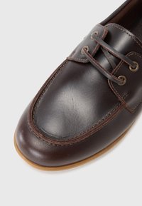 Scarpa da barca in pelle marrone con punta arrotondata, cuciture a contrasto e due occhielli per i lacci. Presenta una suola in gomma per una migliore aderenza.