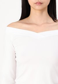 White off-the-shoulder top with a v-shaped neckline and long sleeves, made of smooth, stretchy fabric. Simple design with minimal detailing.