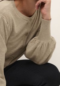 Krémový svetr na knoflíky s balónov�ými rukávy, který je na osobě sedící a odpočívající si na ruce, v kombinaci s černými kalhotami.