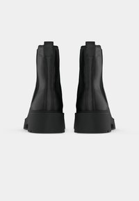 Botas de tornozelo em couro preto com textura suave, apresentando um solado grosso preto e um pequeno salto. As alças estão posicionadas na parte de trás.