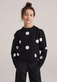 Pull noir en maille avec de gros pois blancs, col rond et coupe décontractée. Associé à un pantalon noir plissé.