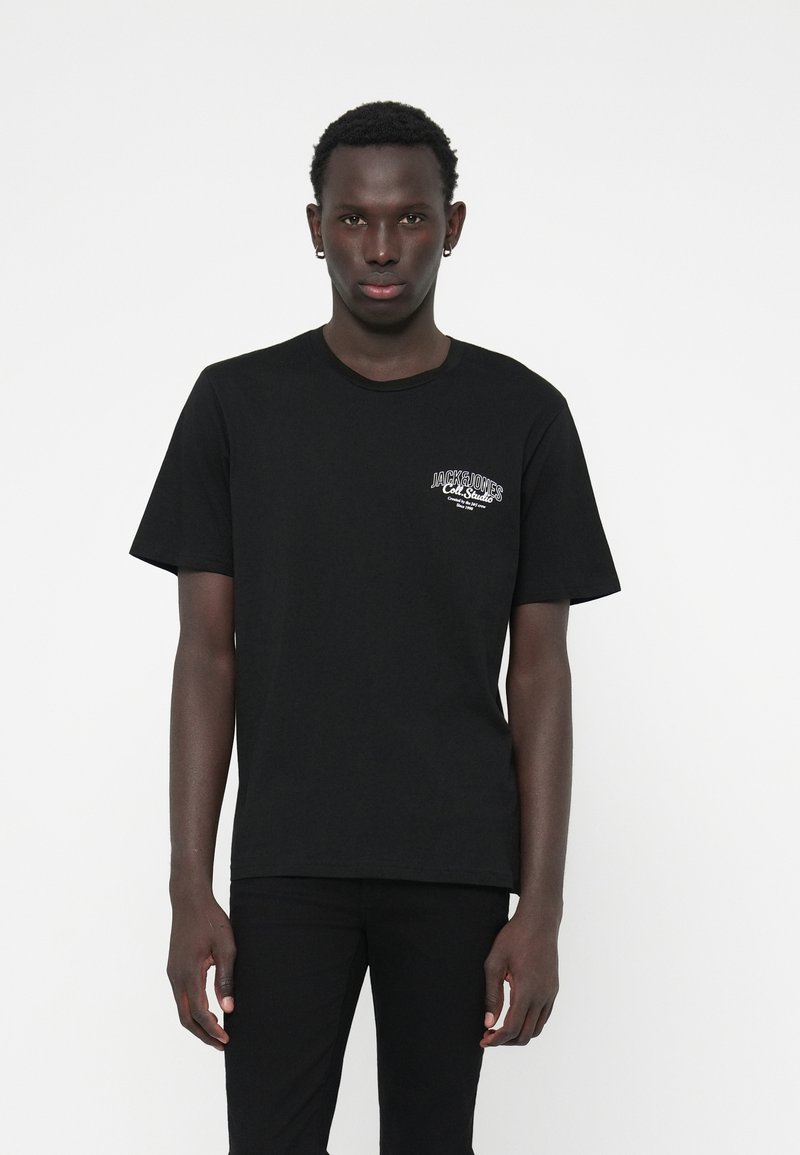 Jeune homme portant un t-shirt noir avec un petit logo blanc "JACK & JONES" sur la poitrine gauche, se tenant contre un fond blanc uni.