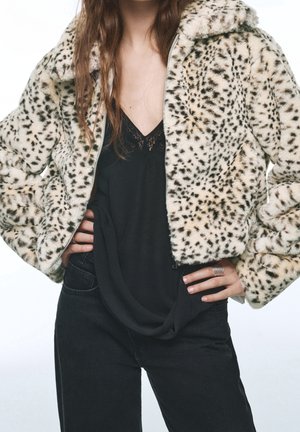 Femme portant une veste courte en fausse fourrure à pois sur un top noir orné de dentelle et un pantalon noir, les mains posées sur les hanches.