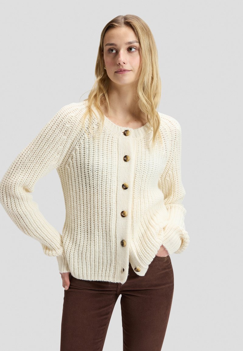 Jeune femme portant un cardigan en maille crème avec de gros boutons et un pantalon marron, debout les mains dans les poches devant un fond uni.