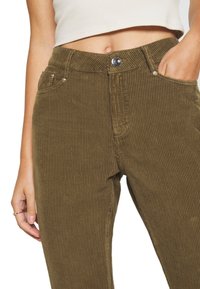 Gros plan sur une personne portant un pantalon en velours côtelé vert olive avec un bouton et une fermeture éclair, la main dans la poche, et un top court en maille blanche.