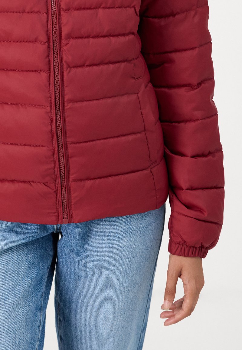 Doudoune bordeaux avec un design matelassé, fermeture éclair et poignets élastiques. Associée à un jean en denim bleu clair. Tissu texturé.