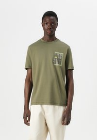 T-shirt en coton vert olive à manches courtes. Présente une poche poitrine avec un design graphique distinctif dans des couleurs atténuées. Ourlet droit.