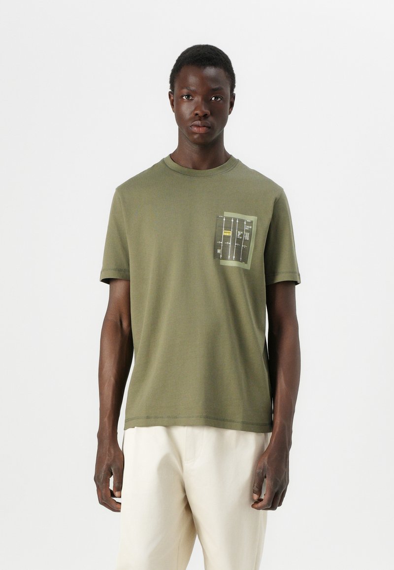 T-shirt en coton vert olive à manches courtes. Présente une poche poitrine avec un design graphique distinctif dans des couleurs atténuées. Ourlet droit.