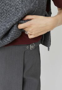Pull en tricot gris superposé sur un haut bordeaux, avec un pantalon gris doté d'un clip argenté et de plis bien taillés. Gros plan d'une main sur le pull.