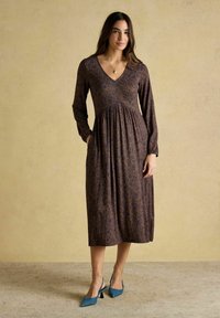 Frau trägt ein langärmeliges, V-Ausschnitt, knielanges Blumenkleid mit Taschen und blaue Absatzschuhe, steht vor einem beigen Hintergrund.