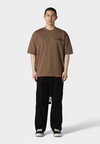 Brun oversized bomulds t-shirt med korte ærmer, med en sort grafik på brystet. Sammen med sorte cargo bukser og sneakers.
