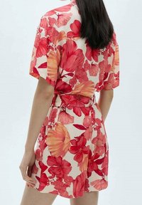 Króca sukienka w kwiaty w kremowym kolorze, z dużymi czerwonymi i pomarańczowymi nadrukami kwiatowymi. Z krótkimi rękawami i wiązanym w pasie.