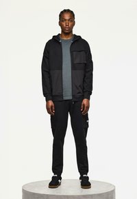 Zwarte zip-up hoodie met gestructureerde voorzakken, gecombineerd met zwarte cargobroeken. Vergezeld van zwarte sneakers met gumrubberen zolen.