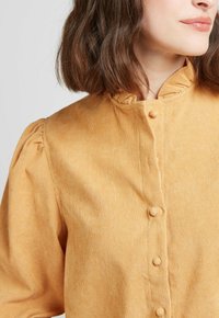 Chemise en velours côtelé jaune moutarde avec col froncé et fermeture à boutons, dotée de manches bouffantes et d'un motif côtelé texturé.