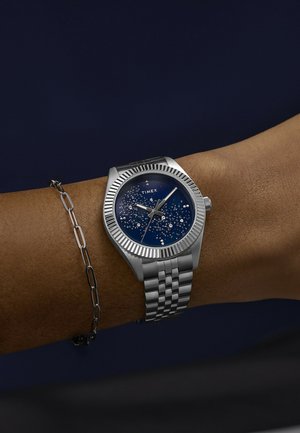 Randmelg hõbedase Timex-kellaga, millel on tumesinine tähine sihverplaat ja hõbedane metallist käevõru, koos hõbedase kettaga.