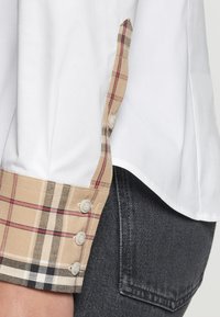 Camicia bianca con cuffie a quadri beige che presentano accenti rossi e neri. La manica ha tre bottoni e una texture liscia. Indossata con jeans scuri.