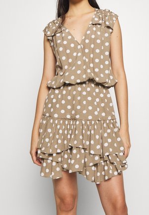 Beżowa sukienka bez rękawów w białe kropki, z dekoltem w serek, falbanką na dole i marszczonym pasem, noszona przez stojącą osobę na jednolitym tle.