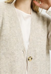 Sweter kardigan w jasnym beżu o miękkiej fakturze, z dekoltem w kształcie v, zapięciem na guziki z przodu i lekkim rozcięciem. Pod spodem widać kremową bluzkę.