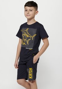 Mėlyna T-shirt su geltonais ryklių grafikais ir taškų raštu; derinama su atitinkamomis šortais, kuriuose matyti "KRSH" geltona spalva ir taškų akcentai.