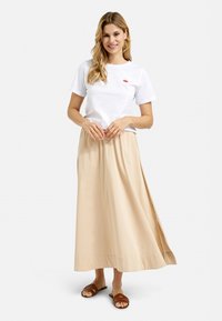  Weißes Baumwoll-T-Shirt mit rotem Krabbenlogo, kombiniert mit einem fließenden beigen Maxirock. Braune Rutschen-Sandalen vervollständigen das Outfit. Einfaches und lässiges Design.