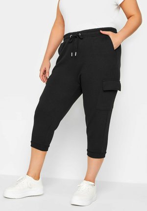 Frau trägt schwarze, gekürzte Cargo-Hosen mit Kordelzugbund, weiße Sneaker und weißes ärmelloses Oberteil, steht mit einer Hand in der Tasche.