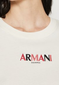 Närbild av en person som bär en vit skjorta med en röd och svart broderad "ARMANI EXCHANGE"-logotyp på bröstet.