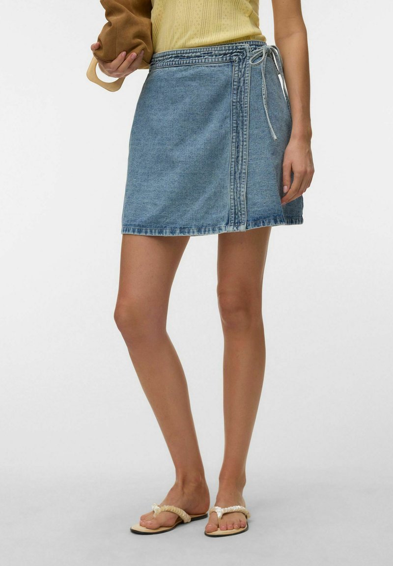 Femme portant une jupe portefeuille en jean longueur genou avec un détail noué, des sandales tressées beige, et tenant un sac marron.
