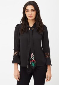 Blusa negra con un lazo en el cuello, bordados florales en rosa y rojo, y detalles de encaje en las mangas. Tejido suave con una forma ajustada.