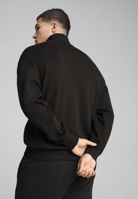 Sort strikpullover med høj krave, afslappet pasform og lange ærmer. Modellen står med hænderne samlet bag ryggen, præsenteret på en neutral baggrund.