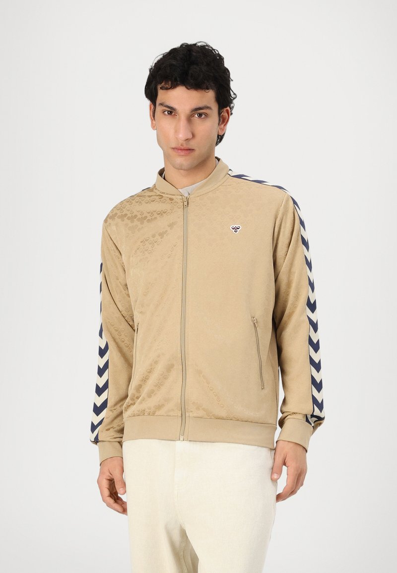 Beige zip-up jacket with a textured pattern, navy chevron stripes on sleeves, and a small logo on the chest. Made of soft material.