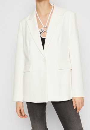 Vrouw die een wit colbert met één knoop draagt over een zwarte top met een witte, bandjesachtige halslijn, gecombineerd met donkergrijze jeans.