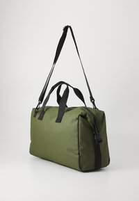 Sac de voyage vert olive avec une texture rembourrée, bandoulière noire ajustable et doubles poignées noires. Compartiment principal zippé visible.