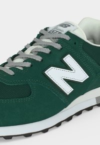 Πράσινο αθλητικό παπούτσι New Balance 574 με γκρι κορδόνια, λευκό λογότυπο "N" και λευκή επενδυμένη σόλα σε λευκό φόντο.