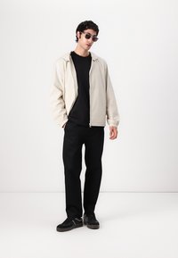 Light beige zip-up jacket with elastic cuffs, worn over a black t-shirt and black pants; paired with dark sunglasses and black shoes.