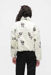 Jakna od kremastog flisa s izvezenim uzorcima smeđih ruža, visokim ovratnikom i skraćeni dizajn. Mekana tekstura i širok kroj s dugim rukavima.