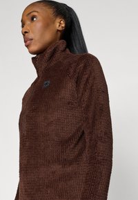 Pullover de fleece castanho com gola alta, apresentando um padrão texturizado, fecho éclair frontal e um pequeno logotipo no peito.