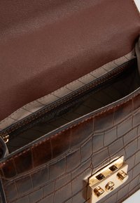 Sac à main en cuir marron texturé avec un intérieur lisse et marron foncé. Dispose d'un fermoir doré et d'une poche zippée, avec un revêtement matelassé.